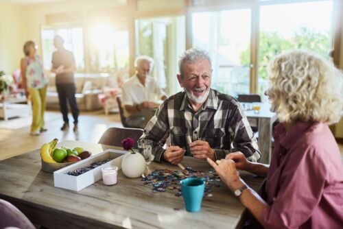 Senior man talking to his friend while assembling a jigsaw puzzle in common room at retirement home. Visual concept for a blog discussing Ohio nursing home staffing mandates.